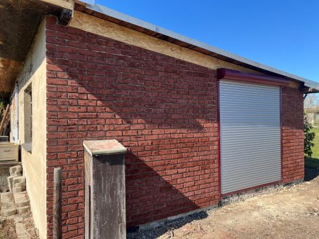 Eckansicht eines Gebäudes mit roten Klinkerriemchen und grauem Rolltor.