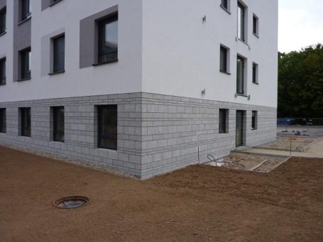 Eckansicht eines modernen, mehrstöckigen Gebäudes mit grauem Sockel und Fenstern.